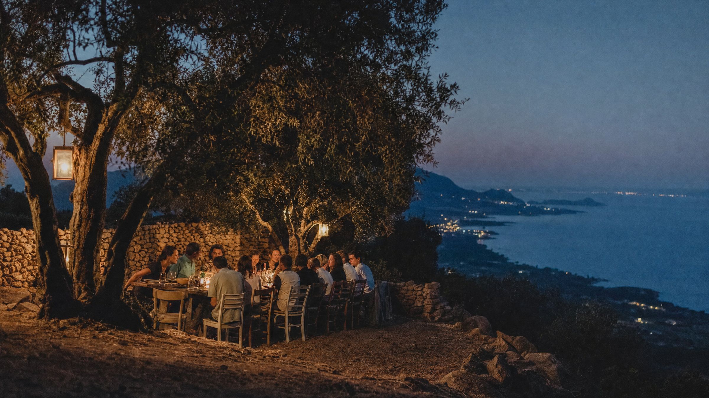 People in conversation at a long table under olive trees on a Sardinian hillside at dusk